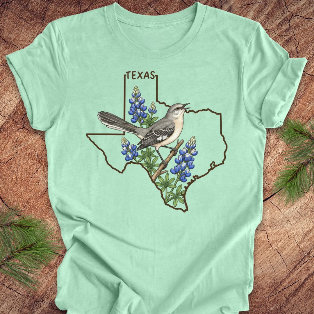 A mint green short-sleeve t-shirt laid flat on a rustic wood background, featuring a graphic of a Texas state outline with a gray bird perched on a branch among bluebonnets, and the word TEXAS above.