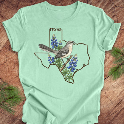 A mint green short-sleeve t-shirt laid flat on a rustic wood background, featuring a graphic of a Texas state outline with a gray bird perched on a branch among bluebonnets, and the word TEXAS above.
