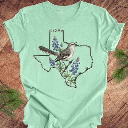 Mint green t-shirt with Texas state outline with state bird, mockingbird and state flower bluebonnet design on a wooden surface
