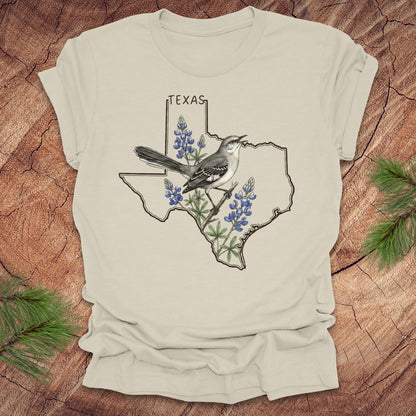 Beige t-shirt with Texas state outline with state bird, mockingbird and state flower bluebonnet design on a wooden surface