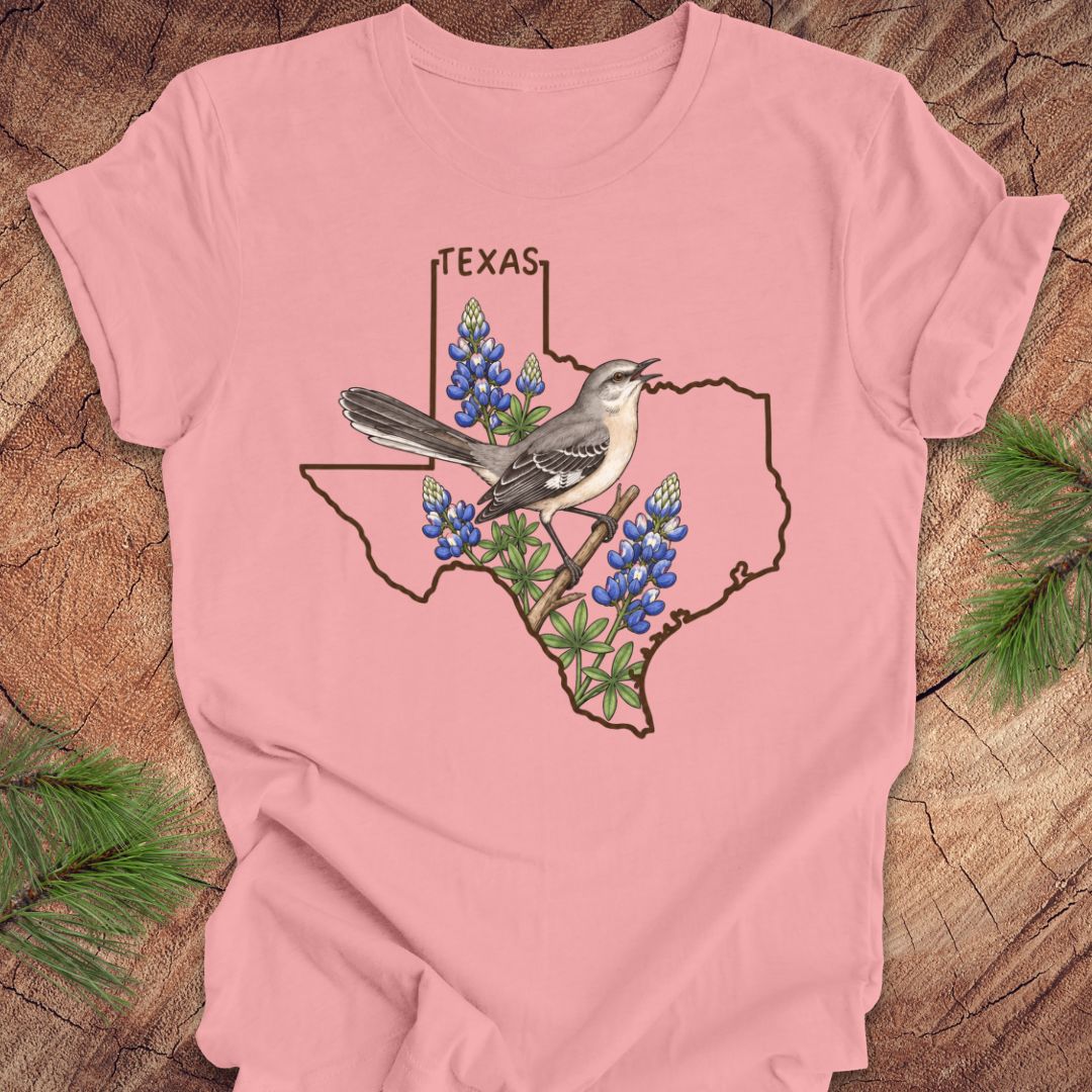 A pink short-sleeve t-shirt with a graphic of a Texas state outline, a mockingbird, and bluebonnets, laid flat on a rustic wood background with pine branches.