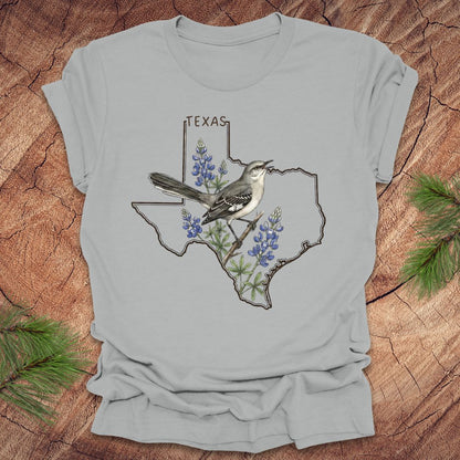 Silver t-shirt with Texas state outline with state bird, mockingbird and state flower bluebonnet design on a wooden surface