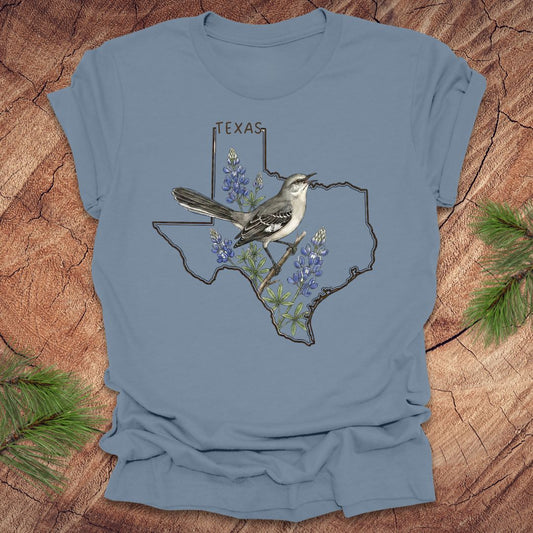 Steel blue t-shirt with Texas state outline with state bird, mockingbird and state flower bluebonnet design on a wooden surface