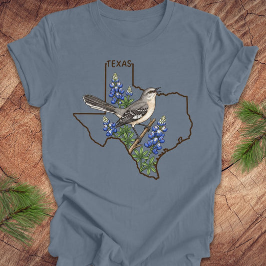 A slate blue short-sleeve t-shirt with a graphic of a gray and white bird perched on a branch over bluebonnets inside the outline of Texas, with the word 'TEXAS' above. The shirt is laid flat on a rustic wood background with pine needles in the corners.