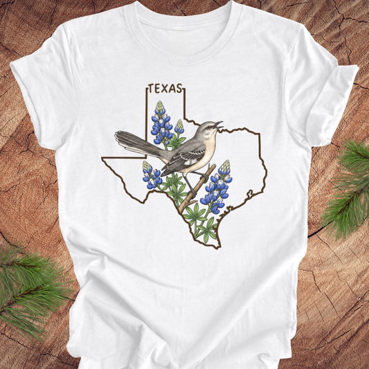A white short-sleeve crewneck t-shirt featuring a graphic of a Texas state outline with a mockingbird perched on bluebonnets and the word 'TEXAS' above, displayed flat on a rustic wood background with pine needles.