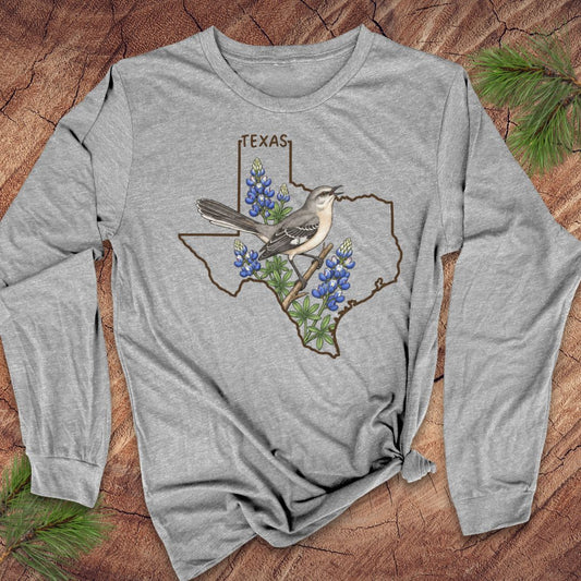 A heather gray long-sleeve t-shirt with a graphic of a Texas state outline, bluebonnets, and a gray bird, laid flat on a rustic wood background with pine needles.