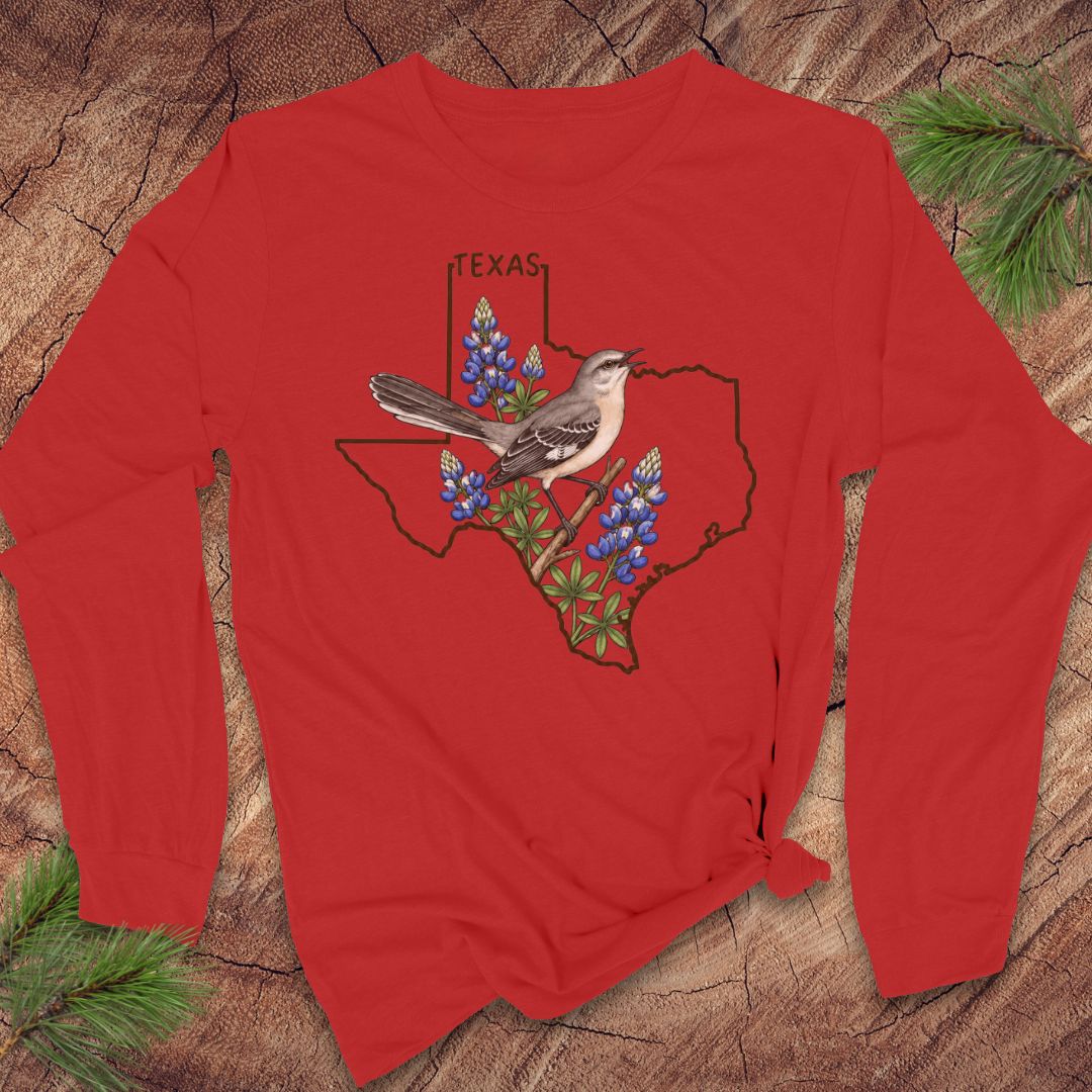 A red long-sleeve t-shirt with a graphic of a Texas state outline, bluebonnets, a gray bird, and the word 'TEXAS' printed on the front, displayed flat on a rustic wood background with pine needles.