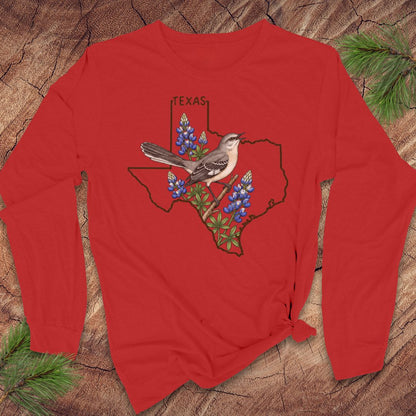 A red long-sleeve t-shirt with a graphic of a Texas state outline, bluebonnets, a gray bird, and the word 'TEXAS' printed on the front, displayed flat on a rustic wood background with pine needles.