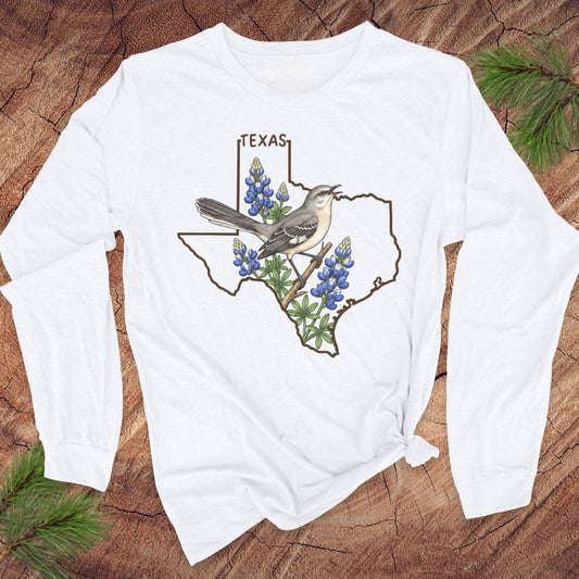 A white long-sleeve t-shirt with a graphic of a Texas state outline, a mockingbird, and bluebonnets, displayed flat on a rustic wood background with pine branches.