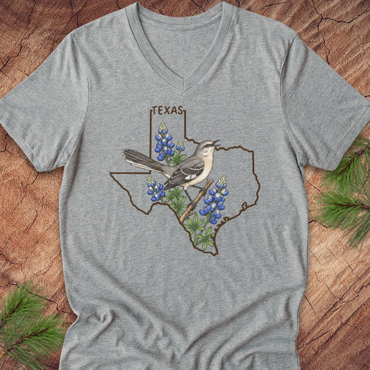 A heather gray V-neck t-shirt featuring a graphic of the state outline of Texas with a gray bird perched on bluebonnets and the word 'TEXAS' above, displayed flat on a rustic wood background with pine branches.