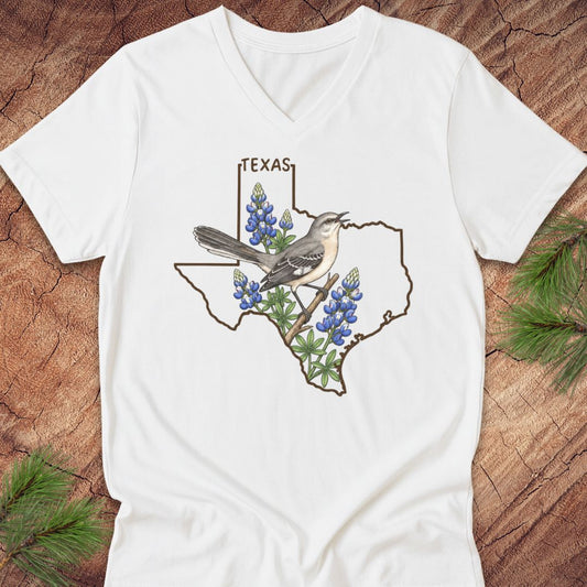 A white V-neck t-shirt laid flat on a rustic wood background with pine branches in the corners. The shirt features a graphic of the state outline of Texas with bluebonnets and a gray and white bird in the center, with the word 'TEXAS' above.