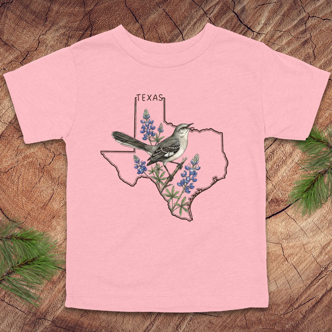Pink toddler t-shirt with Texas state outline and mockingbird on a wood background.