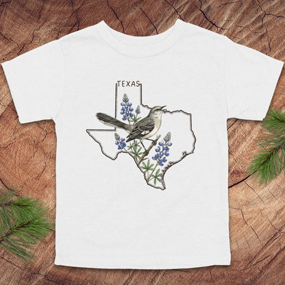 White toddler t-shirt with Texas state outline and mockingbird on a wood background.