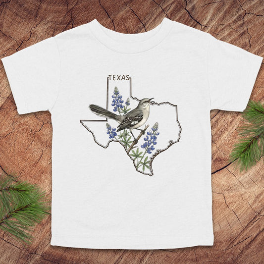 White toddler t-shirt with Texas state outline and mockingbird on a wood background.