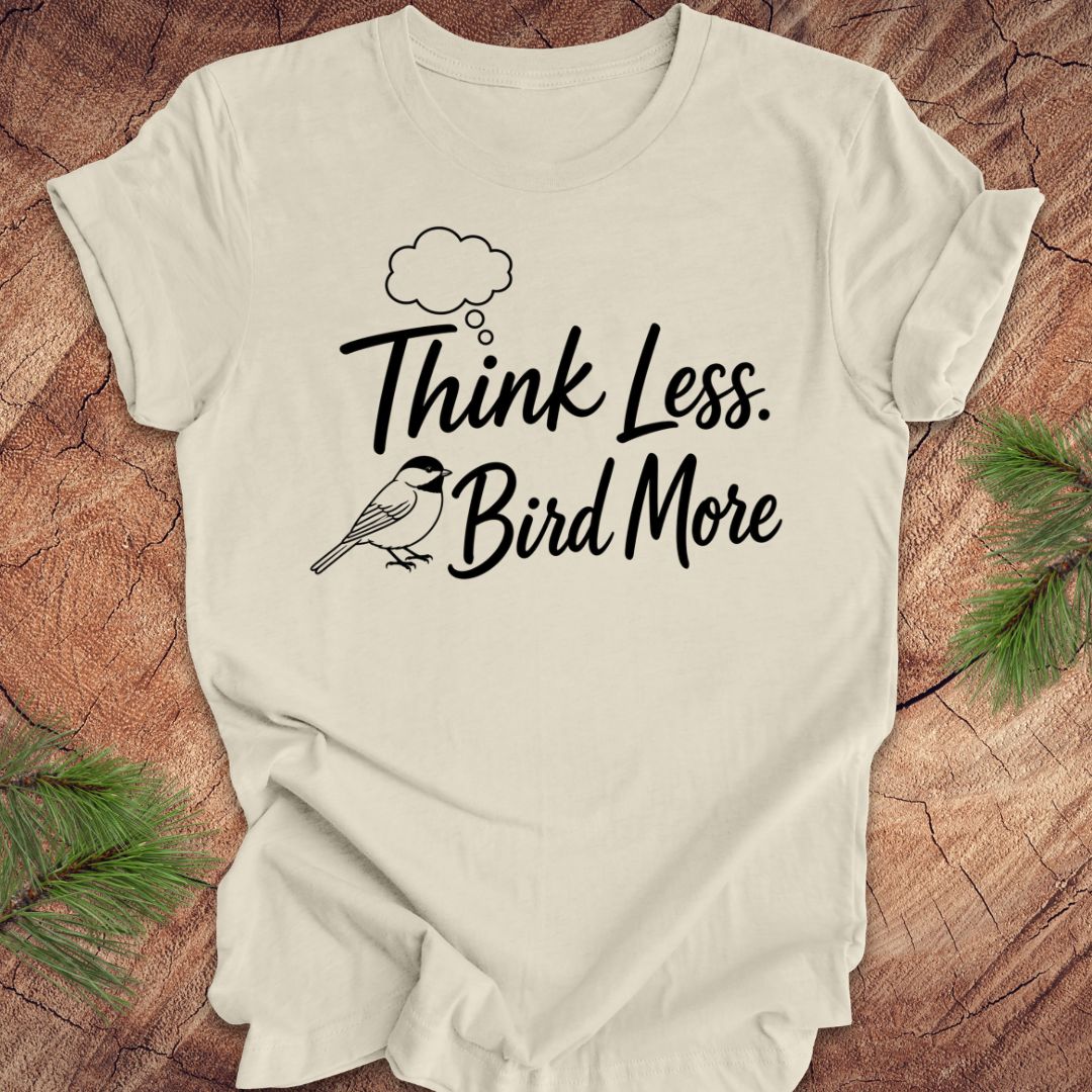 Beige t-shirt with 'Think Less. Bird More' text on a wooden surface with greenery.