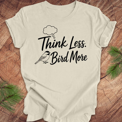 Beige t-shirt with 'Think Less. Bird More' text on a wooden surface with greenery.