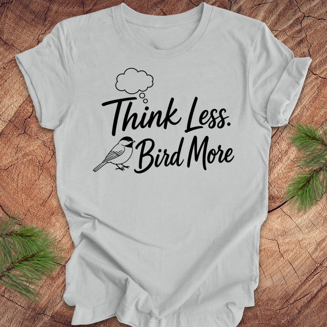 Gray t-shirt with 'Think Less. Bird More' text on a wooden surface with greenery.