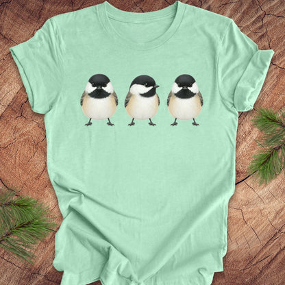 Light green t-shirt with three chickadee illustrations on a wooden surface.