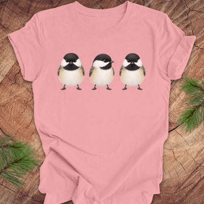 Pink t-shirt with three chickadee illustrations on a wooden surface with pine branches.