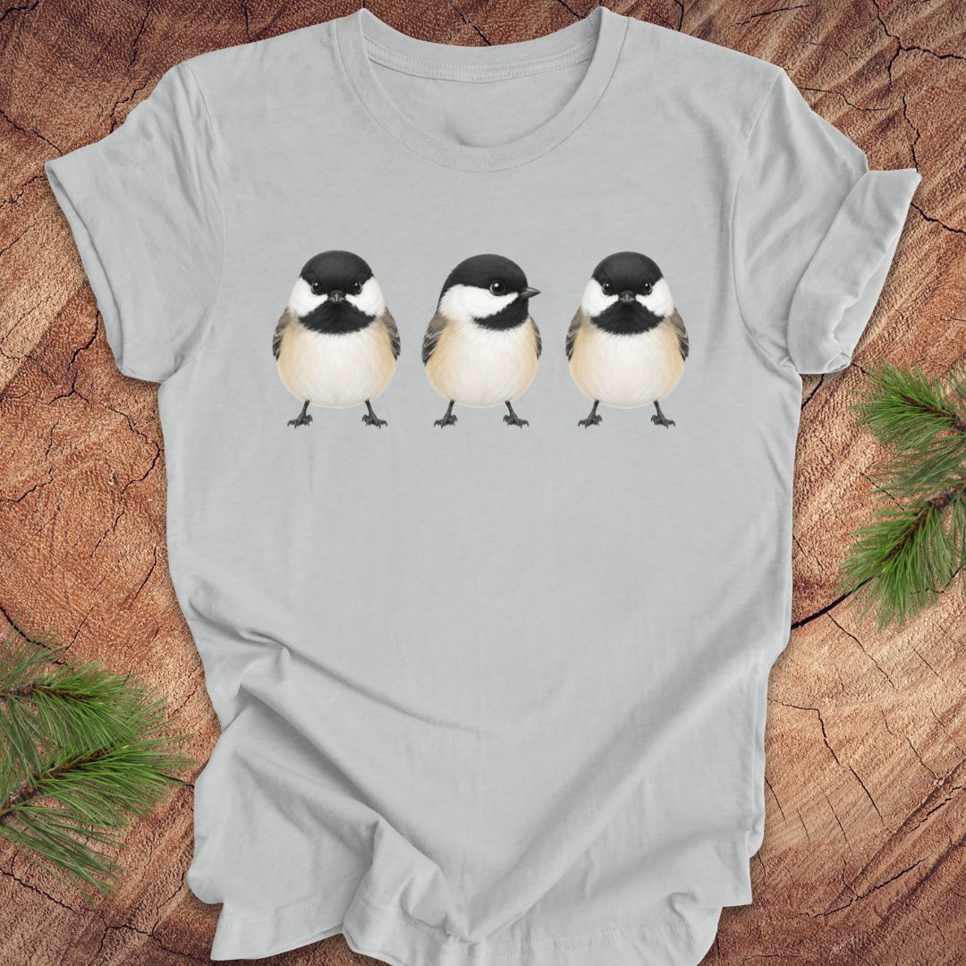 Gray t-shirt with three chickadee illustrations on a wooden surface with pine branches.