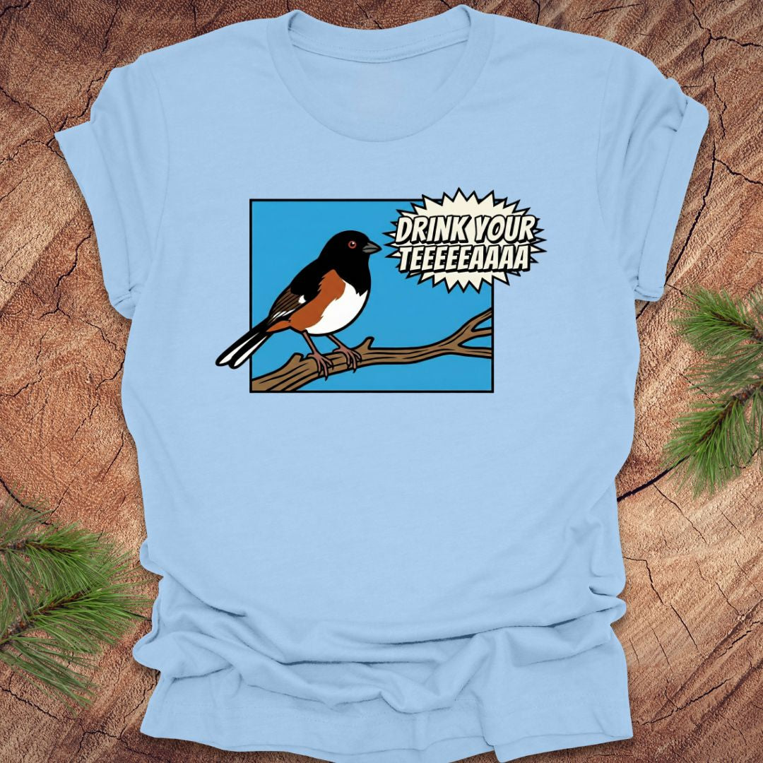 Baby blue t-shirt with a cartoon towhee bird graphic and text, 'drink your teeeaaa" on a wooden surface.