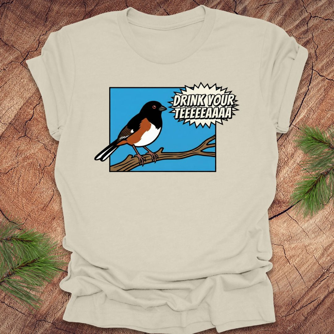 Beige t-shirt with a cartoon towhee bird graphic and text, 'drink your teeeaaa" on a wooden surface.