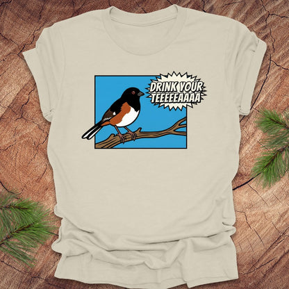 Beige t-shirt with a cartoon towhee bird graphic and text, 'drink your teeeaaa" on a wooden surface.