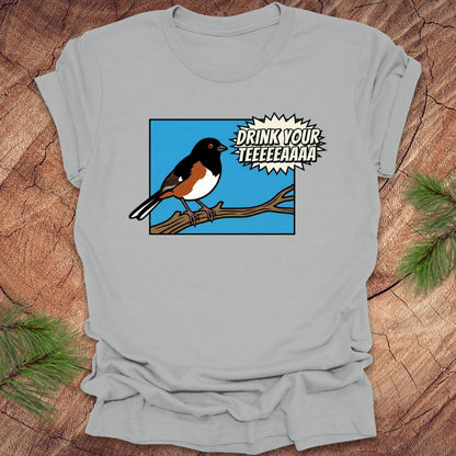 Silver t-shirt with a cartoon towhee bird graphic and text, 'drink your teeeaaa" on a wooden surface.