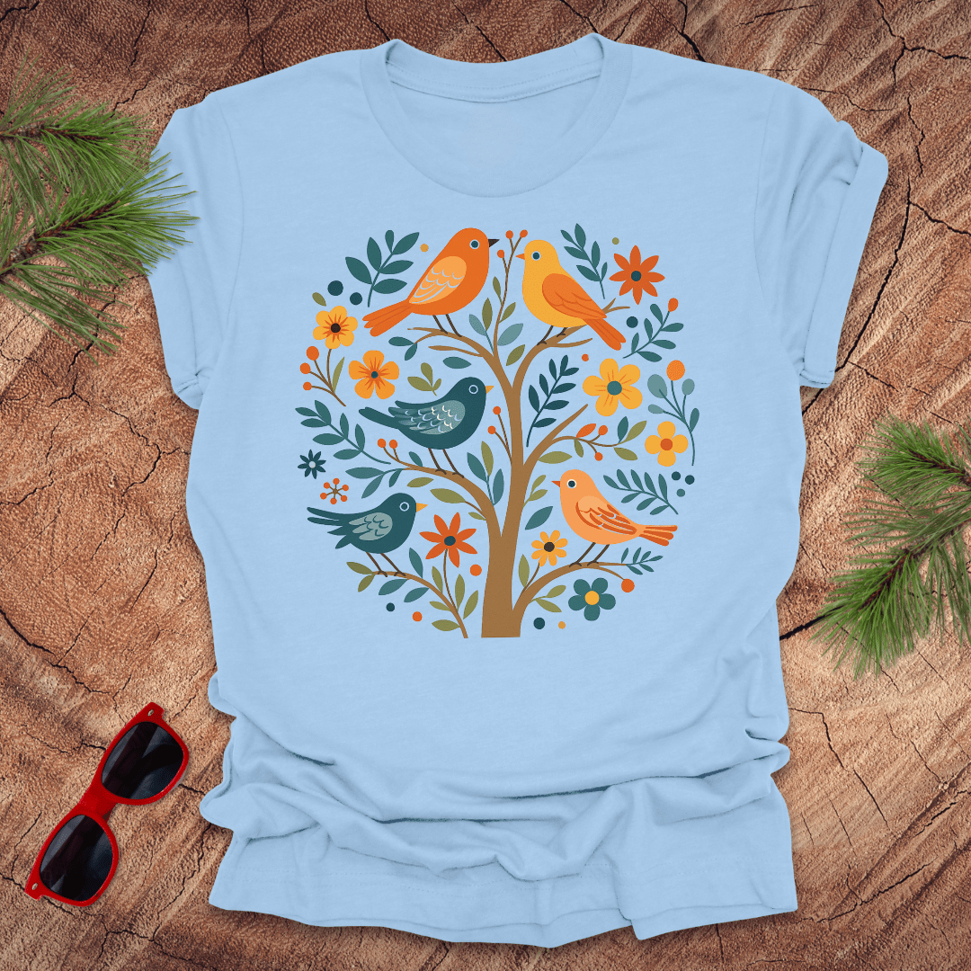 Light blue t-shirt with a colorful bird and floral design on a wooden surface with sunglasses and greenery.