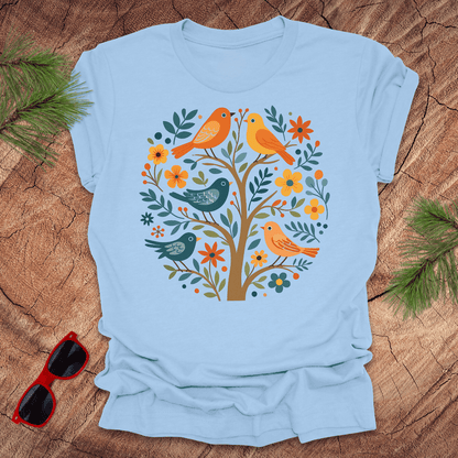 Light blue t-shirt with a colorful bird and floral design on a wooden surface with sunglasses and greenery.