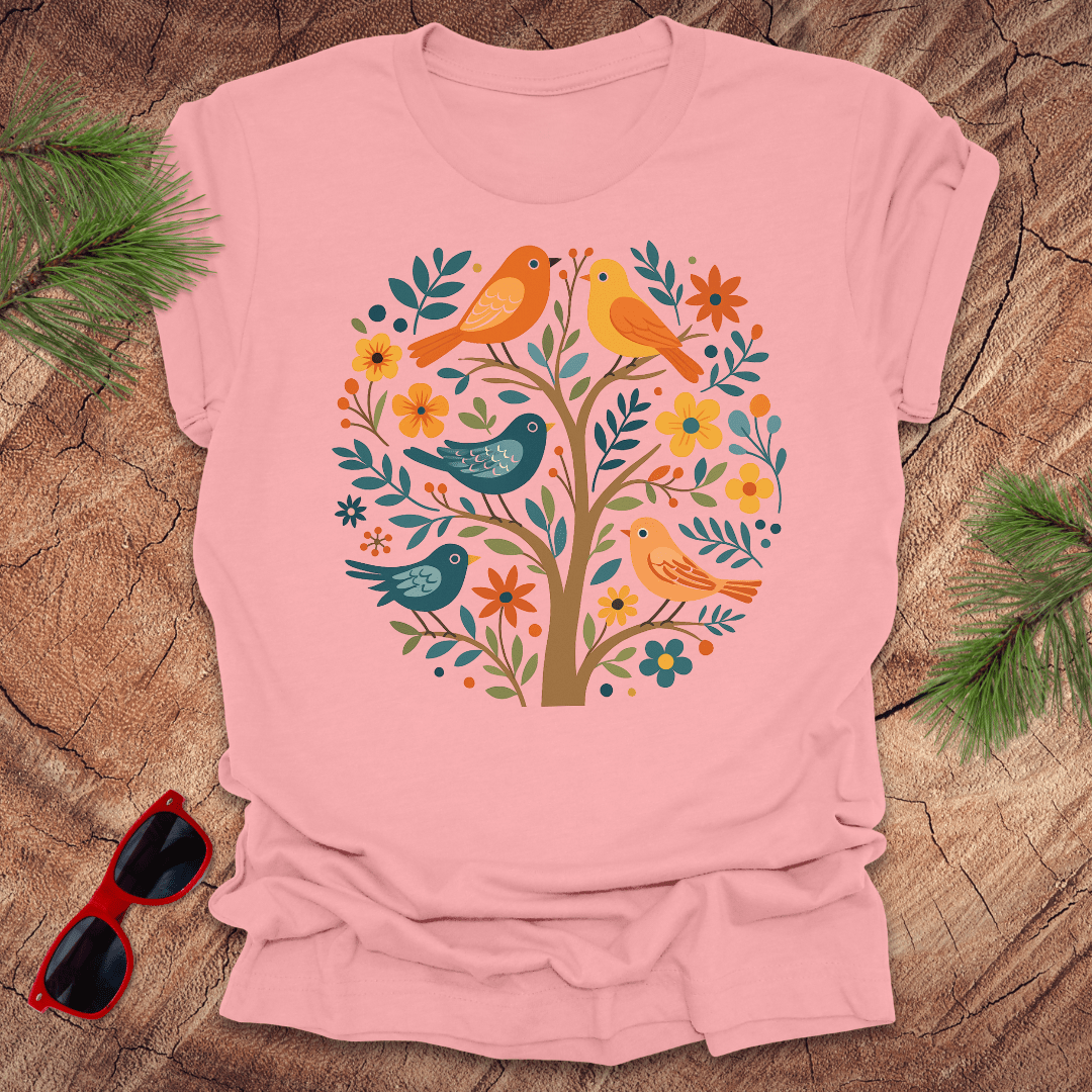 Pink t-shirt with a colorful bird and floral design on a wooden surface with sunglasses and greenery.