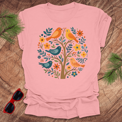 Pink t-shirt with a colorful bird and floral design on a wooden surface with sunglasses and greenery.