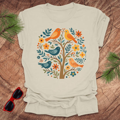 Beige t-shirt with a colorful bird and floral design on a wooden surface with sunglasses and greenery.