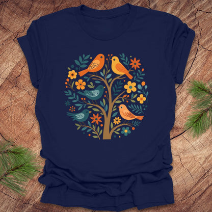 Navy blue t-shirt with colorful bird and tree design on a wooden surface with sunglasses and pine branches.