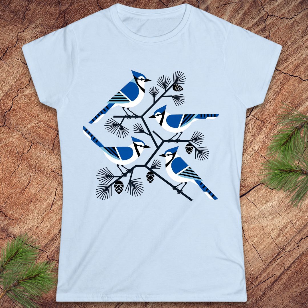A light blue short-sleeve crewneck t-shirt featuring a graphic print of four blue jays perched on pine branches with pinecones, laid flat on a rustic wood background with green pine needles in the corners.