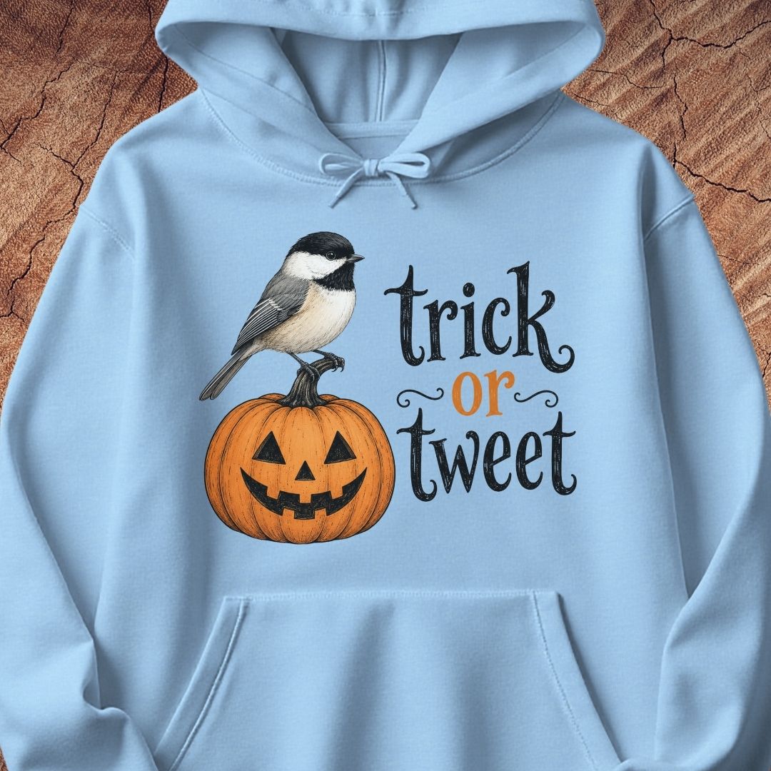 A light blue hoodie laid flat on a textured brown wood background, featuring a graphic print of a black-capped chickadee perched on a carved orange pumpkin next to the text 'trick or tweet' in black and orange lettering.