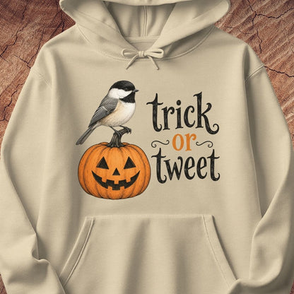 A beige hoodie featuring a graphic print of a chickadee bird perched on a carved orange pumpkin next to the text 'trick or tweet' in black and orange lettering, displayed against a rustic wood background.
