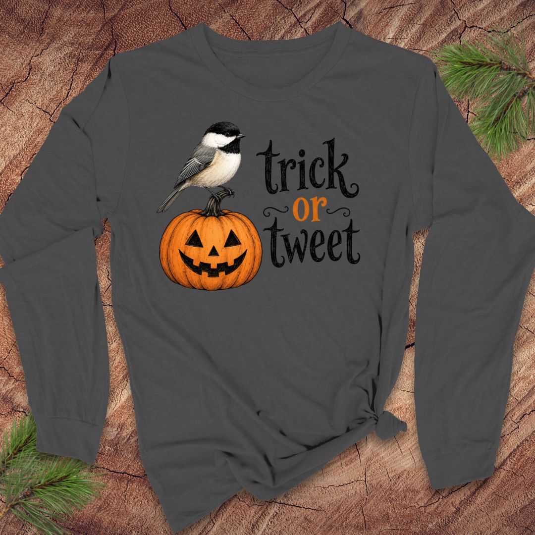 A dark gray long-sleeve t-shirt with a Halloween graphic featuring a chickadee bird on a jack-o'-lantern pumpkin and the text 'trick or tweet' in black and orange, displayed flat on a rustic wood background with pine branches.
