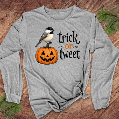 A heather gray long-sleeve t-shirt with a Halloween graphic featuring a chickadee on a jack-o'-lantern and the text 'trick or tweet'. The shirt is laid flat on a rustic wood background with pine needles.