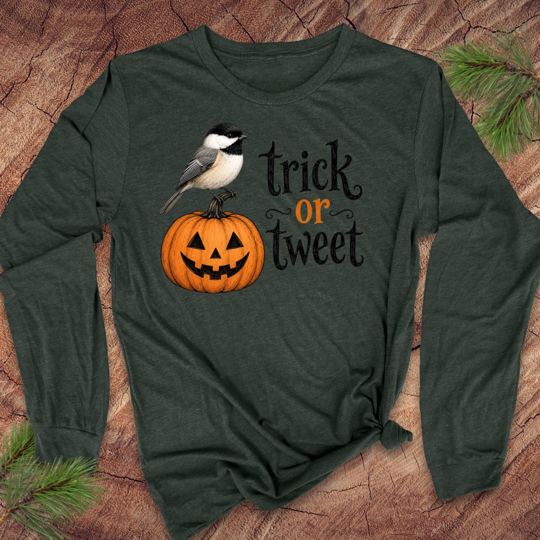A dark forest green long-sleeve t-shirt with a 'trick or tweet' Halloween graphic, laid flat on a rustic wood background with pine needles. The graphic features a black-capped chickadee perched on a carved orange pumpkin next to the text 'trick or tweet' in black and orange.