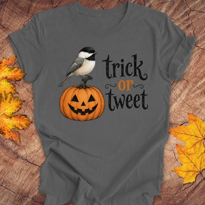 A dark gray graphic t-shirt featuring a cute design of a black-capped chickadee bird perched on a jack-o'-lantern pumpkin next to the text 'trick or treat' in black and orange lettering. The t-shirt is displayed flat on a rustic wood background decorated with autumn leaves.