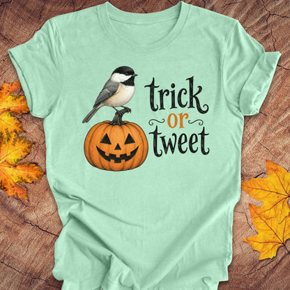 A mint green graphic t-shirt laid flat on a rustic wood background with autumn leaves. The shirt features a cartoon illustration of a black-tipped chickadee perched on a carved orange jack-o'-lantern pumpkin, next to the text 'trick or tweet' in black and orange lettering.