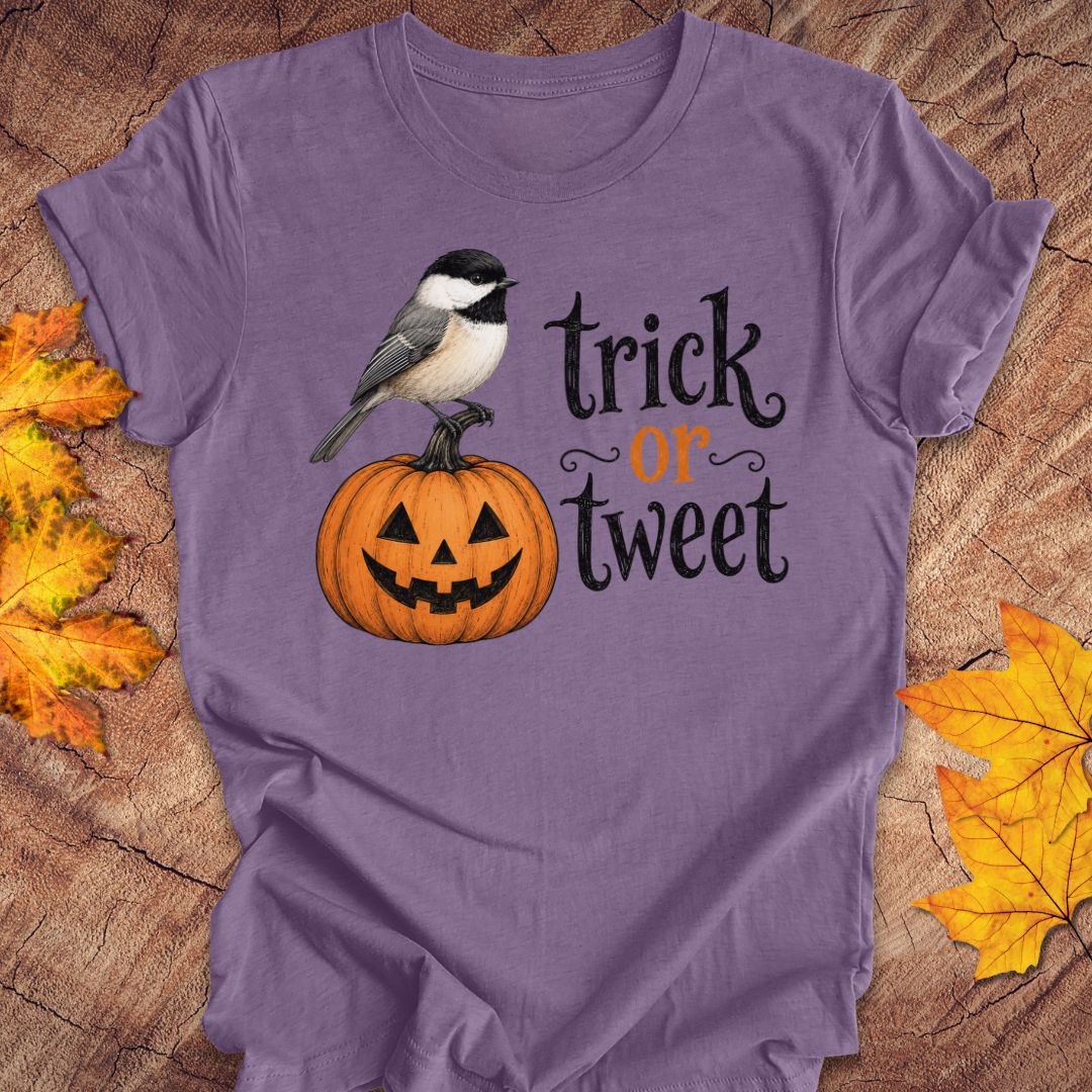 A purple short-sleeve T-shirt laid flat on a wood surface with autumn leaves around it. The shirt has a printed graphic of a small black-and-white bird perched on an orange jack-o'-lantern next to the words 'trick or treat' in black and orange lettering.