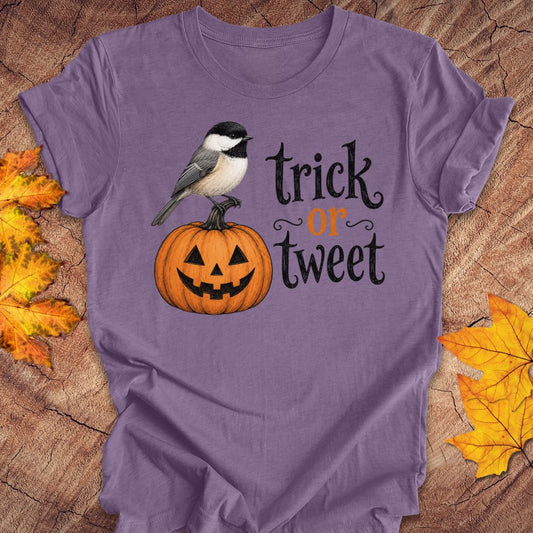 A purple short-sleeve T-shirt laid flat on a wood surface with autumn leaves around it. The shirt has a printed graphic of a small black-and-white bird perched on an orange jack-o'-lantern next to the words 'trick or treat' in black and orange lettering.