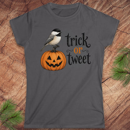 A dark gray short-sleeve crewneck t-shirt featuring a graphic of a black-capped chickadee perched on a carved orange jack-o'-lantern pumpkin next to the text 'trick or tweet' in black and orange lettering. The t-shirt is displayed flat on a rustic wood background with pine needles in the corners.