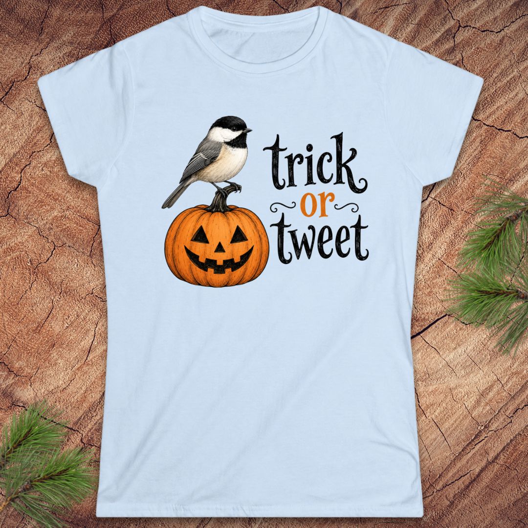 A light blue short-sleeve crewneck t-shirt featuring a graphic of a black-capped chickadee perched on a carved orange pumpkin next to the text 'trick or tweet' in black and orange lettering. The t-shirt is displayed flat on a rustic wood background with pine needles in the corners.