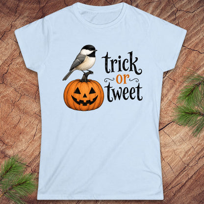 A light blue short-sleeve crewneck t-shirt featuring a graphic of a black-capped chickadee perched on a carved orange pumpkin next to the text 'trick or tweet' in black and orange lettering. The t-shirt is displayed flat on a rustic wood background with pine needles in the corners.