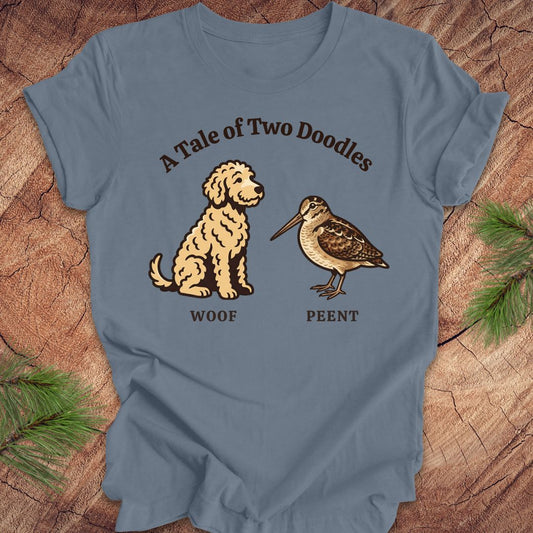 Blue t-shirt with a doodle dog and a woodcock bird with text, 'a tale of two doodles' on a wood background.