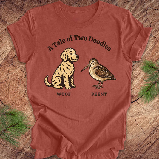 Clay colored t-shirt with a doodle dog and a woodcock bird with text, 'a tale of two doodles' on a wood background.