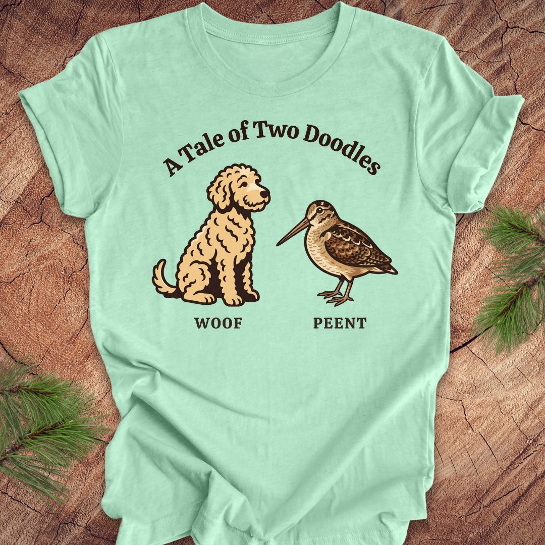Mint green t-shirt with a doodle dog and a woodcock bird with text, 'a tale of two doodles' on a wood background.